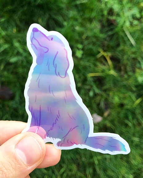 A hand holds a Golden Retriever sticker in front of some greenery