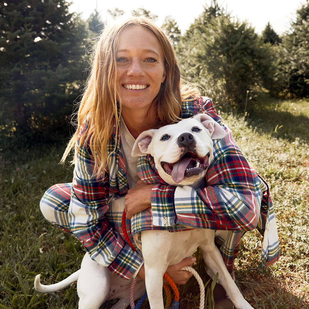 A female Aerie model in plaid holiday pyjamas smiles and hugs a white pitbull