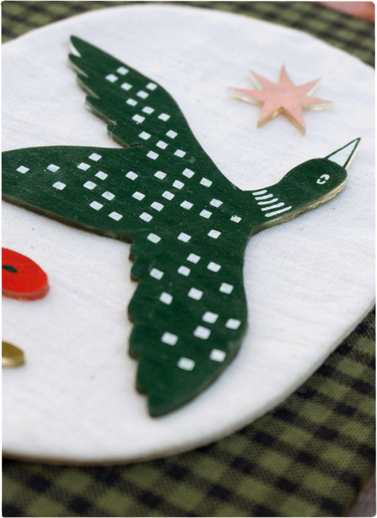 A mixed media artwork of a loon flying between a star and a poppy. It sits on a beige linen oval on top of a green gingham fabric background